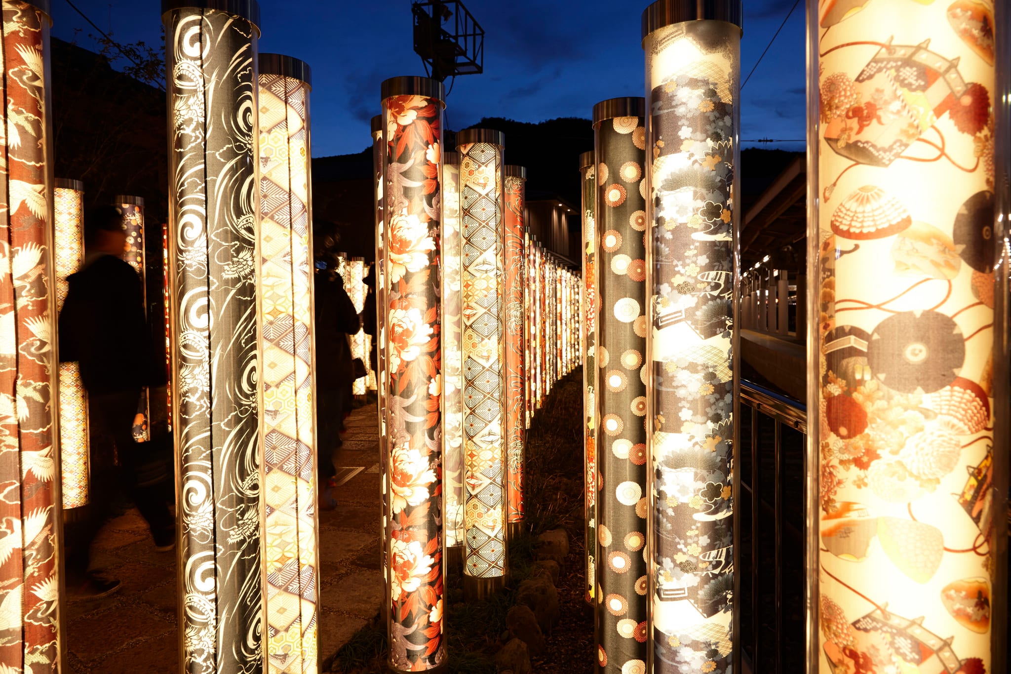 Tall pillars of kimono silk lit from within at night in the Kimono Forest, Arashiyama, Kyoto